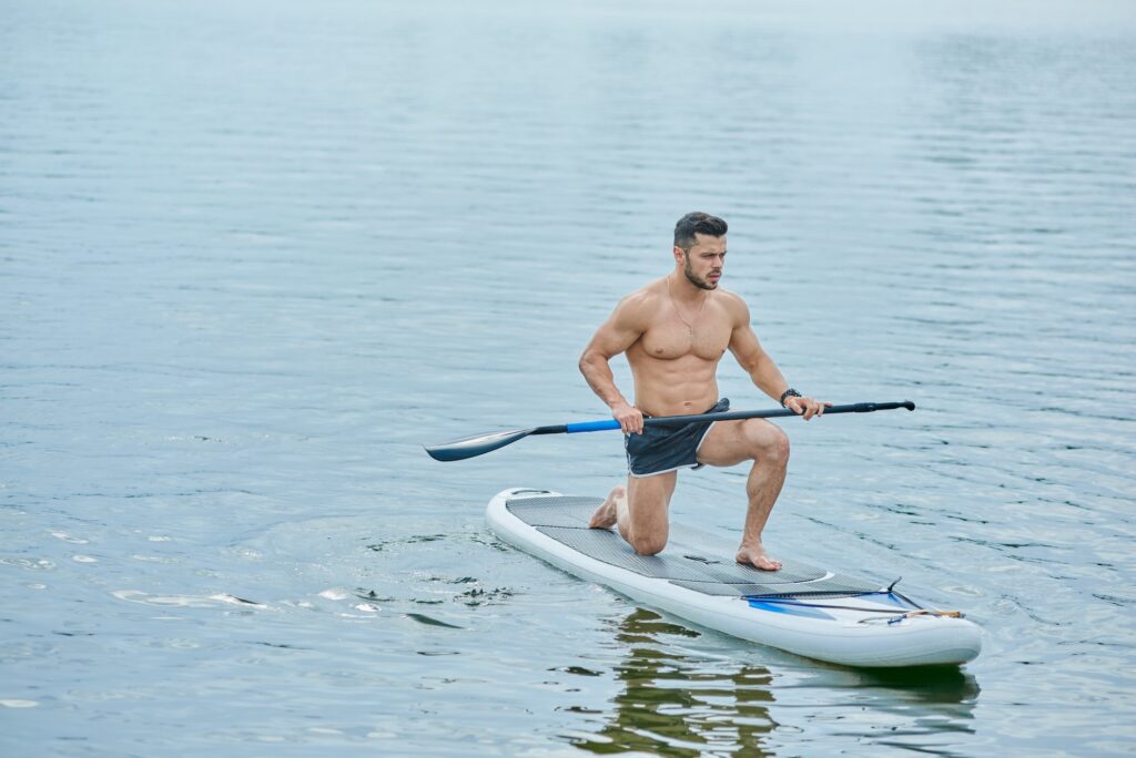 Young sportsman keeping long oar, standing on one one knee, swimming on sup board in city lake